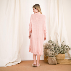 Una mujer de espaldas viste un vestido largo de color rosa claro, con mangas largas y un diseño suelto, en un entorno luminoso con decoraciones naturales.