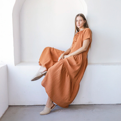 Una mujer sentada en una pared blanca, vistiendo un vestido largo color terracota con mangas cortas y un lazo en el escote.