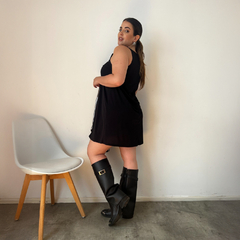 Modelo usando un vestido negro corto llamado Vestido Isabella, de pie junto a una silla blanca, con botas altas negras.