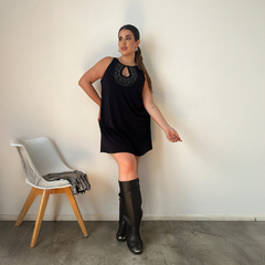 Modelo luciendo un vestido negro con detalles brillantes en el escote, posando junto a una silla con accesorios.