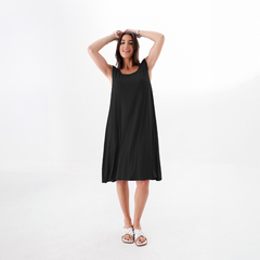 Una mujer sonriente lleva un vestido negro de corte suelto, con sandalias blancas, en un fondo blanco.