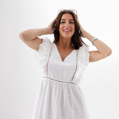 Modelo usando un vestido blanco con mangas de volantes y detalles bordados, sonriendo y sosteniendo gafas en la cabeza.