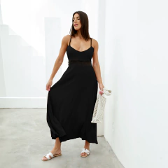 Modelo luciendo un vestido largo negro con tirantes finos y un diseño elegante, sosteniendo una bolsa de red, en un entorno minimalista.