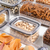 Pote hermético retangular transparente contendo sementes, cercado por outros potes com diferentes alimentos como macarrão e grãos.