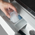 Uma mão segurando um recipiente transparente com líquido, prestes a colocá-lo em um dispenser de lavanderia.