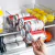 Organizador multiuso porta latas com latas de refrigerante e frutas em uma geladeira.
