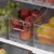 Três organizadores de geladeira transparentes contendo uvas, maçãs e laranjas, dispostos em uma prateleira de refrigerador.