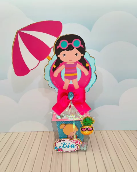 Centro de mesa decorativo com tema de festa pool party, apresentando uma menina com boia, óculos de sol e um guarda-sol, adornado com laço rosa e detalhes coloridos.