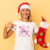 Modelo sorridente usando uma camiseta com estampa natalina, segurando uma meia de Natal, em fundo amarelo.