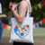 Uma mulher segurando uma bolsa de tecido branca com ilustrações de materiais escolares, celebrando o Dia dos Professores.