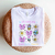 Camiseta branca com estampa colorida sobre a profissão de professora, incluindo desenhos de flores, laços, um globo terrestre e frases motivacionais.