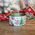 Lata decorativa de Natal com a inscrição "Feliz Natal", adornada com elementos festivos como árvores e enfeites.