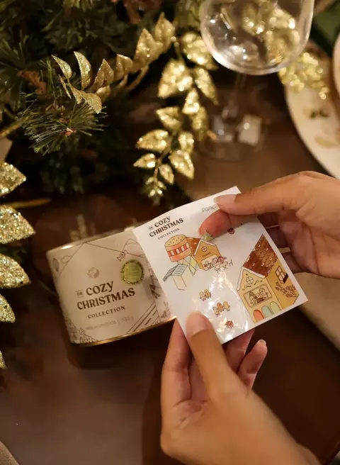 Mãos segurando uma cartela de adesivos natalinos da coleção The Cozy Christmas ao lado de uma embalagem de panettone decorada com folhas douradas e taça de vinho