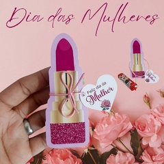 Embalagem decorativa em forma de batom rosa com tag "Feliz dia da Mulher", cercada por rosas.