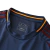 nova terceira camisa da AS Roma da Adidas. A camisa é predominantemente azul marinho e apresenta um escudo retrô circular com as siglas "ASR" no centro. Detalhes em bordô e amarelo, como as três listras nos ombros, recortes nas laterais e a numeração nas 