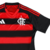 Camisa Flamengo I 2025/26 Feminina Adidas, listrada em vermelho e preto, gola V preta com friso vermelho, ombros pretos com três listras vermelhas da Adidas, escudo CRF branco no peito, logo Adidas branco, numeração e patrocínios em branco nas costas, des