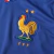 Camisa titular da seleção francesa de futebol 2024-2025. Camisa na cor azul royal com gola V tricolor e o galo, símbolo da seleção, em tamanho maior que o convencional. Na parte interna da gola, a frase "Nos différences nous unissent" (Nossas diferenças n