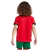 Conjunto Infantil Titular da Seleção Portuguesa Nike 2024-2025. Camisa vermelha com gola preta e verde, calção verde e escudo da FPF. Inspirada na bandeira portuguesa. Vista frontal de um menino vestindo o conjunto completo, em pose de comemoração.