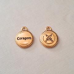 Pingente em formato redondo com a palavra "Coragem" e ícone de espadas cruzadas, em fundo dourado.