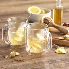 Duas canecas de vidro duplo com bebida quente e pedaços de gengibre, ao fundo limões e um recipiente com óleo.