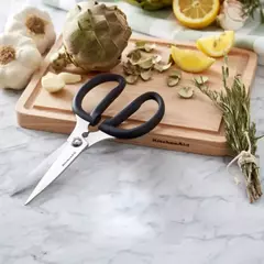 Tesoura de cozinha multiuso preta KitchenAid em uma tábua de corte com alho, limão e ervas ao fundo.