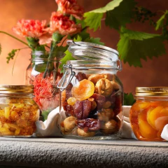 Pote de vidro hermético de 750ml com frutas secas ao centro, cercado por potes menores e flores decorativas ao fundo.