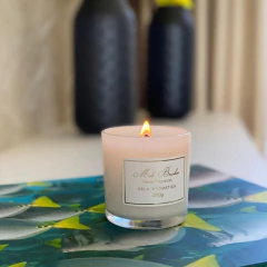 Vela aromática Café Brasil da Mels Brushes acesa, em um suporte transparente, sobre uma mesa com um fundo colorido.