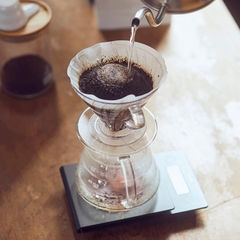 Suporte acrílico Hario V60-03 sendo utilizado para coar café, com água quente sendo vertida sobre o pó de café em um filtro.