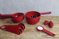 Conjunto de 5 colheres medidoras vermelhas KitchenAid ao lado de uma tigela com frutas e açúcar.