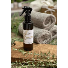 Frasco âmbar com home spray de ambientes e água de tecidos aroma alecrim da marca Mels Brushes ambientado com toalhas de banho ao fundo e ramos de alecrim. 