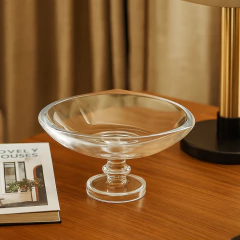 Centro de mesa de vidro transparente com pé, formato quadrado arredondado, sobre mesa de madeira