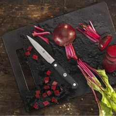 faca para legumes com cabo sintético preto e lâmina gravada com a marca em cima de uma tábua de corte ao lado de uma cebola roxa