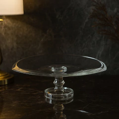 Centro de mesa de vidro transparente com pé, formato redondo e bordas levemente onduladas, sobre superfície escura