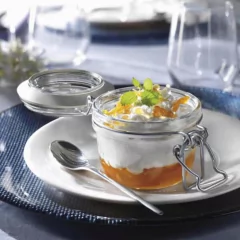 Pote hermético de vidro com sobremesa de frutas e chantilly, decorado com folhas de hortelã, sobre prato branco.