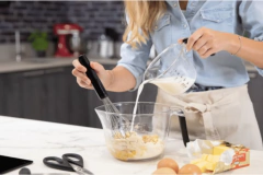 Mulher despejando leite em uma tigela com ingredientes, usando uma jarra medidora KitchenAid de 2 litros.