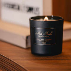 Vela aromática Alecrim 200g da Mels Brushes acesa em um suporte preto, posicionada sobre uma mesa de madeira.