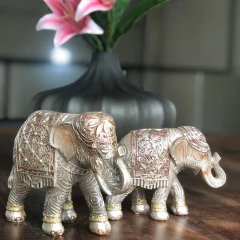 Estatueta de elefantes em prata com detalhes ornamentais, medindo 14x6x10cm, decorando uma mesa com um vaso ao fundo.