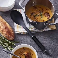Conjunto de utensílios de cozinha KitchenAid com colher de servir ao lado de uma panela com sopa e pão ao fundo.