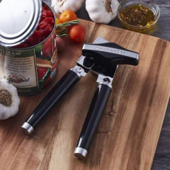 Utensílio de cozinha da marca KitchenAid, um abridor de latas preto, posicionado sobre uma tábua de madeira, com tomates, alho e azeite ao fundo.