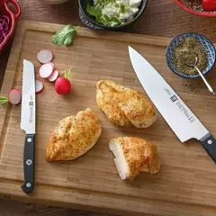 Faca Universal Zwilling Gourmet sobre uma tábua de madeira com pedaços de frango assado e legumes frescos ao redor.
