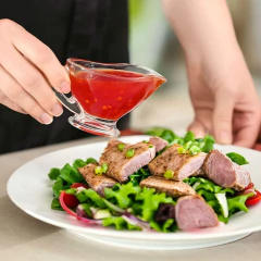 Uma mão segurando uma mini molheira de vidro com molho vermelho, sobre uma salada com folhas verdes e pedaços de carne grelhada.