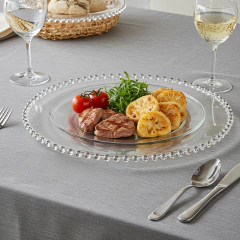 Sousplat cristal com borda de pontos, apresentando um prato com carne grelhada, batatas e salada em uma mesa decorada.