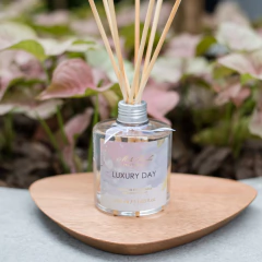 Difusor de aromas Luxury Day de 350ml com varetas em um suporte de madeira, cercado por plantas verdes.