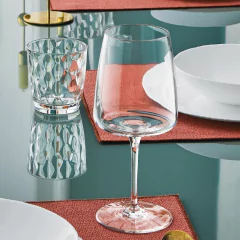 Taça de vinho transparente de 555ml da Bormioli Rocco em uma mesa elegante, ao lado de um copo e pratos brancos.