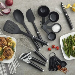 Conjunto de utensílios de cozinha KitchenAid, incluindo colheres, espátula, medidores e acessórios sobre uma superfície de pedra, com pratos de frango e legumes ao lado.