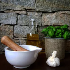 Um pilão de cerâmica branco com um socador de madeira de carvalho, alho e uma garrafa de óleo sobre uma mesa