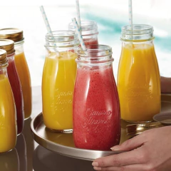 Garrafa de vidro Quattro Stagioni com bebidas coloridas, incluindo sucos e smoothies, em uma bandeja. Há canudos decorativos nas garrafas.