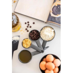 Conjunto de 4 xícaras medidoras cinzas da KitchenAid com ingredientes como chocolate, farinha e ovos sobre uma mesa de cozinha.