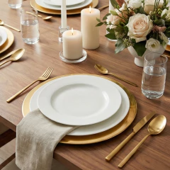 Mesa posta com sousplats dourados de metal, pratos brancos, talheres dourados, velas acesas e arranjo floral