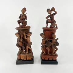 Par de esculturas em madeira representando figuras femininas indianas do século XIX, com detalhes esculpidos de pessoas tocando instrumentos musicais, montadas em base preta.
